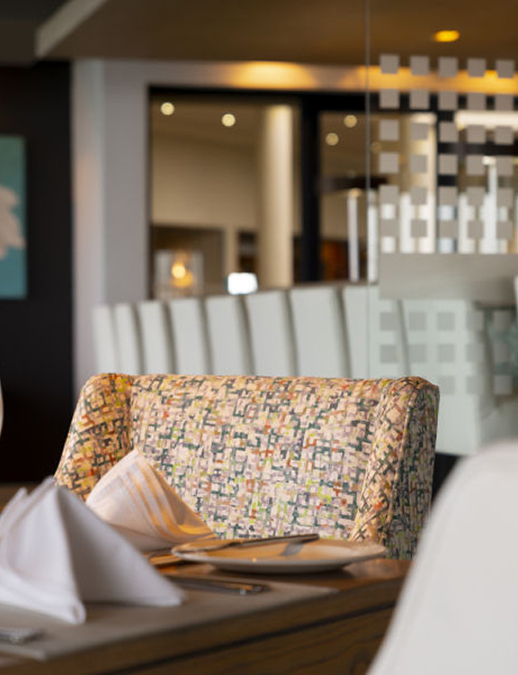 Interior of a restaurant showing a patterned booth, white napkins on a table with cutlery and wine glasses.